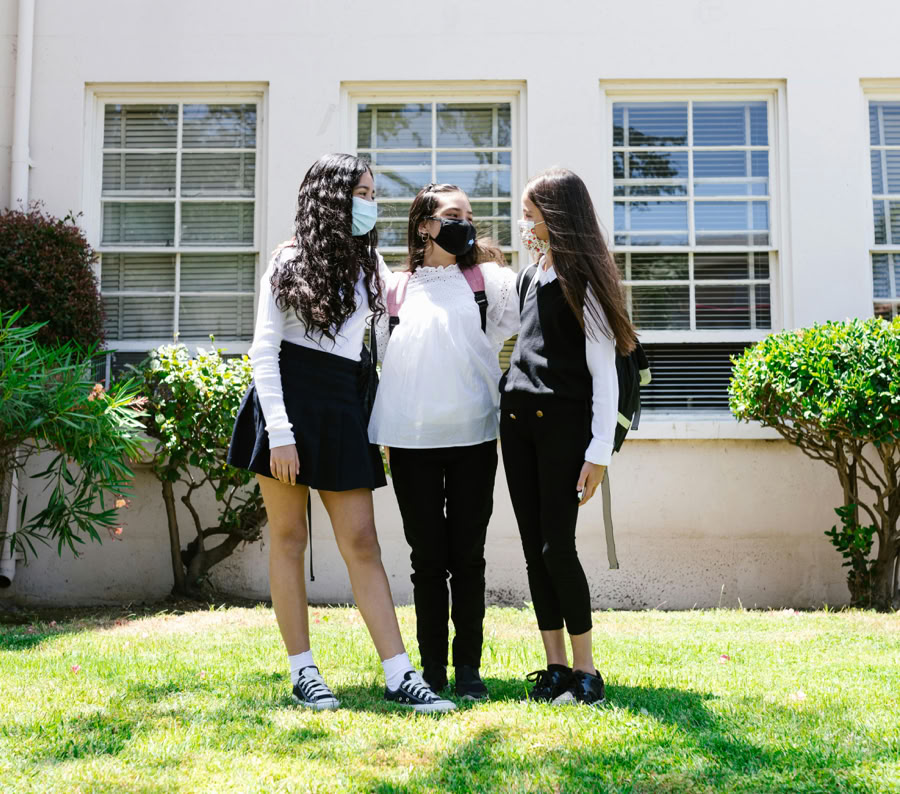 Three teenage girls wearing face masks standing outside a school building, embracing each other.